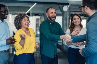 Group of colleagues laughing in an office while two men shake hands