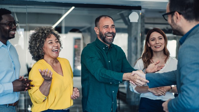 Group of colleagues laughing in an office while two men shake hands