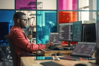 A person working intently at a desk with multiple monitors displaying code and data analysis, in an office with colorful glass partitions.