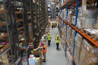 Workers wearing safety vests and helmets inside a large warehouse with tall shelves stocked with various items.