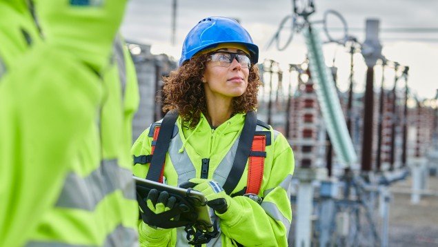 Worker in safety gear, including a blue helmet and reflective vest, holds a tablet and inspects equipment at an industrial site.