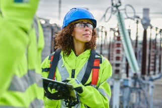 Worker in safety gear, including a blue helmet and reflective vest, holds a tablet and inspects equipment at an industrial site.