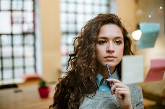 A person contemplates while holding a pen near their chin, looking at sticky notes on a glass wall in a brightly lit office setting.