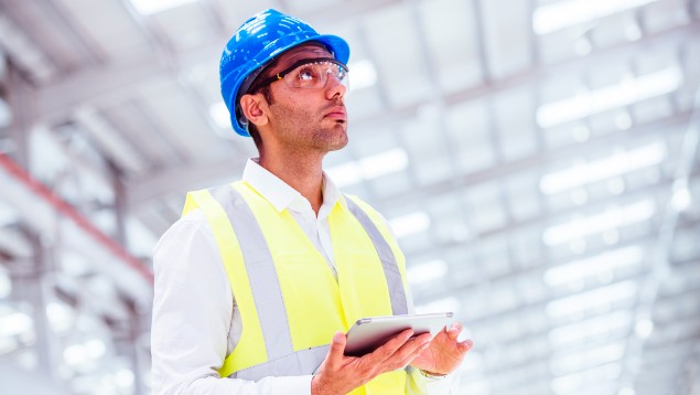 A safety professional in a hard hat and reflective vest holds a tablet while intently observing a factory setting.